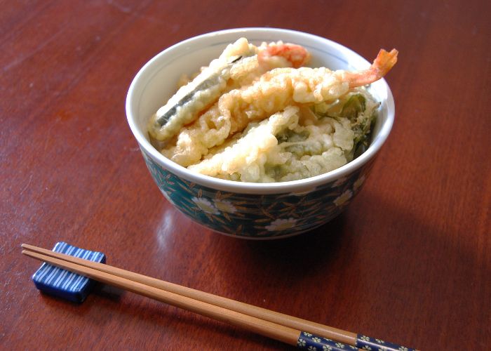 A small bowl of white rice topped with tempura with a pair of chopsticks