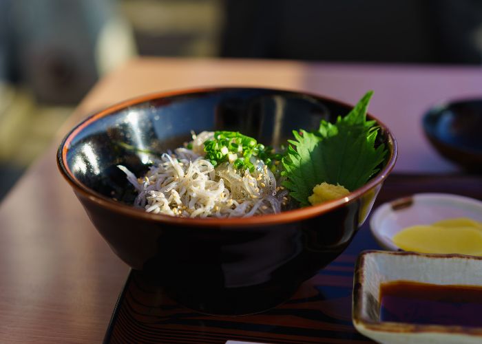 A black wooden bowl of shirasudon, topped with very small white fish, green onions and green herbs