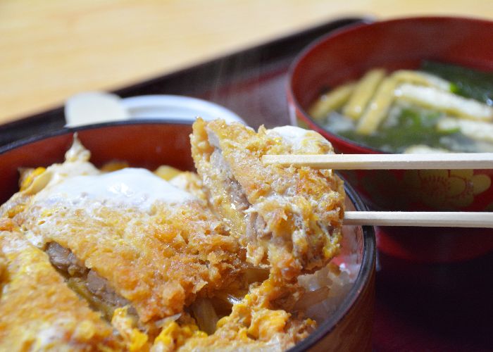 A close up image of a bowl of katsudon, with a pair of wooden chopsticks holding a slice of breaded cutlet