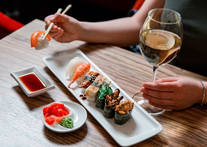 A women holds a glass of wine in one hand and picks up sushi using chopsticks in her other. 