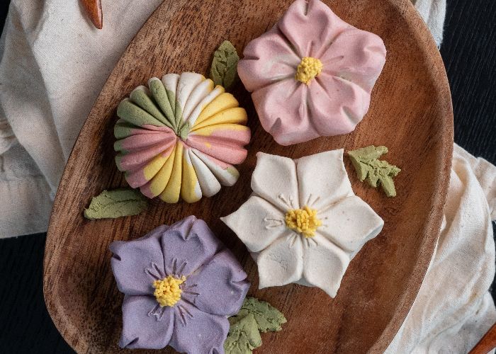 Four wagashi sweets on a wooden plate: a pink flower, a white flower, a purple flower, and a multicolored swirl