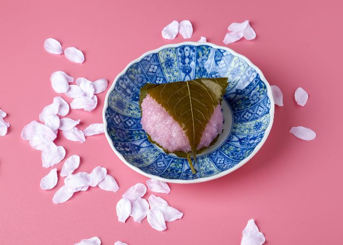 A pink sakura mochi wrapped in a green leaf, on a blue plate against a pink background surrounded by cherry blossom petals