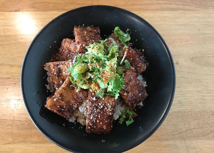 A black bowl of white rice topped with several slices of dark brown grilled tofu and sliced green onions