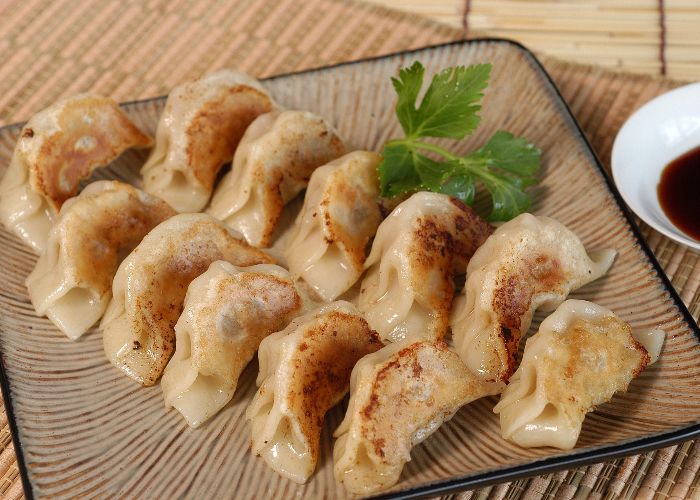 A square plate of 12 small grilled gyoza with a green herb on top and dipping sauce next to it