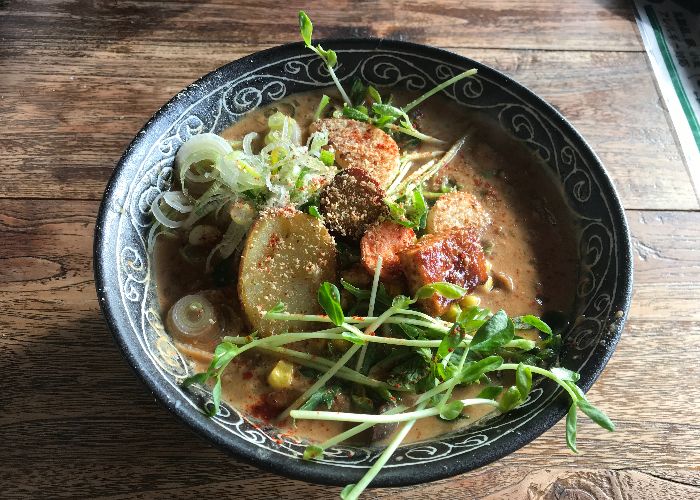 An overhead image of a bowl of ramen, topped with tofu, sliced potatoes, herbs, and sliced green onions, with a brown broth