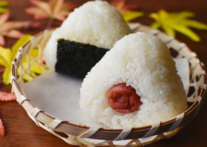 Two triangular onigiri white rice balls in a basket, one wrapped in a sheet of nori seaweed and the other with a ume pickled plum in the center