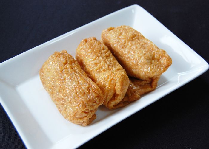 Three light brown inarizushi tofu rice pockets lined up on a rectangular white dish