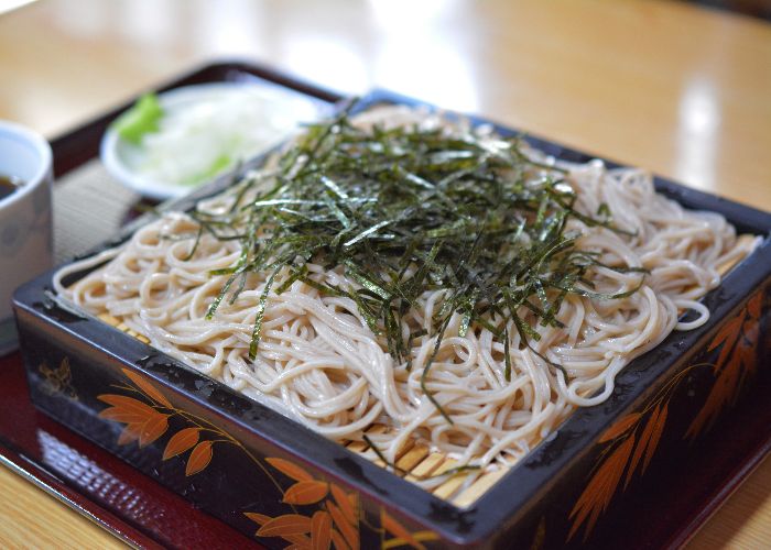 A heap of brown soba noodles in a square lacquered box, topped with thin slices of dark green seaweed