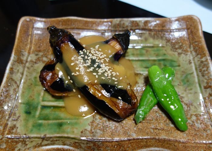 A small grilled eggplant sliced in half and coated with a light brown miso sauce and white sesame seeds, with edamame next to it on the plate