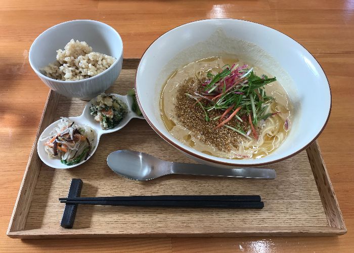 A tray with a a bowl of ramen in a light brown broth topped with vegetables and sesame seeds, a bowl of rice, and a triple dish of little condiments 