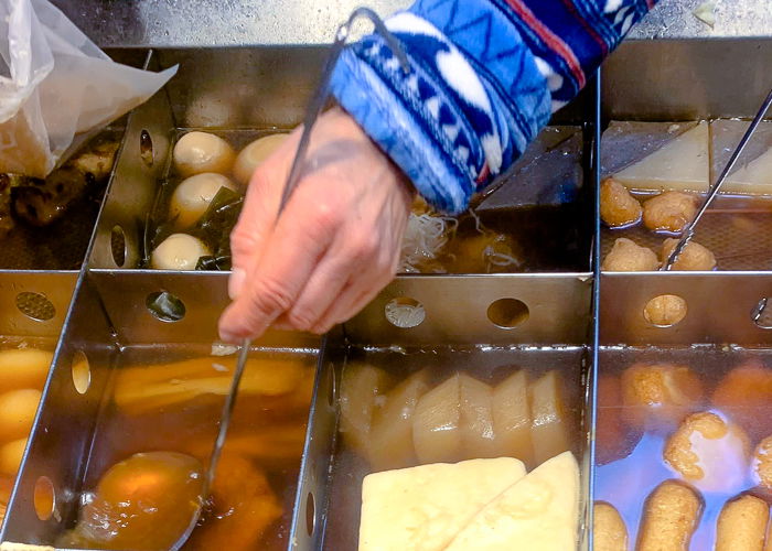 A hand holding a ladle scoops oden soup broth