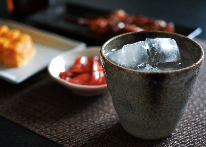 A ceramic grey cup with no handles, filled with clear shochu over ice. In the background there are two small plates of food