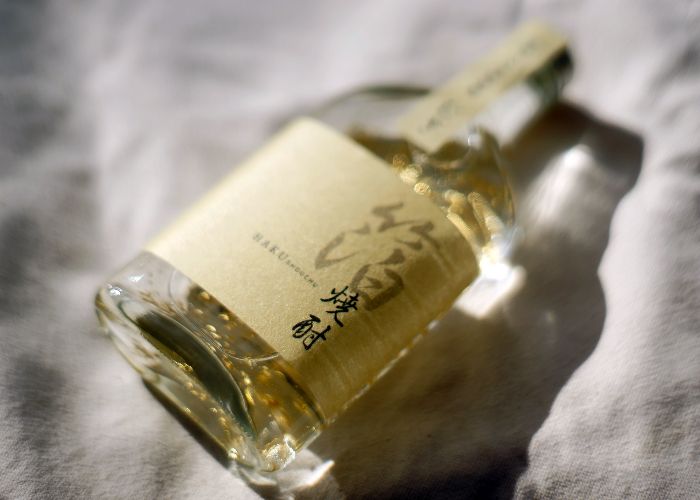 A glass bottle of golden shochu with a gold label, lying on its side on a white cloth backdrop