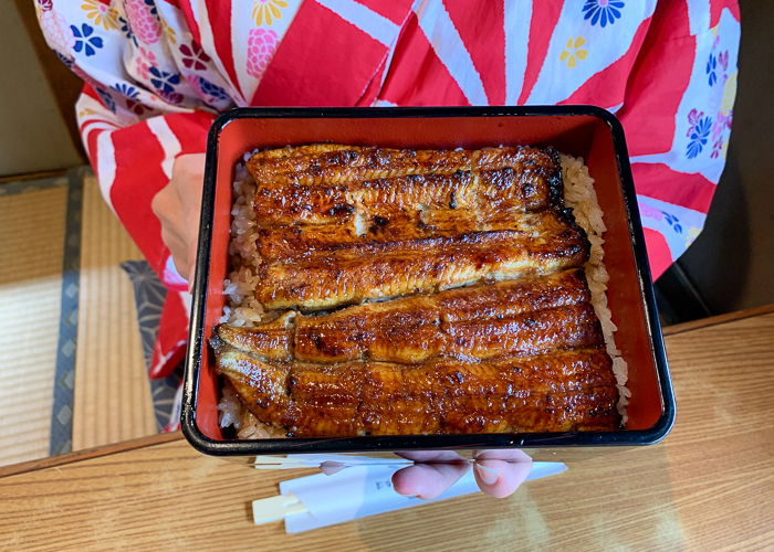 A women in kimono holds a box of unagi over rice