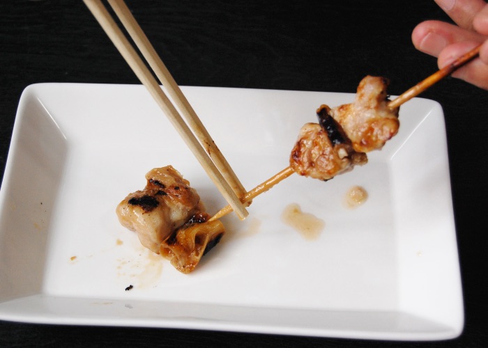 A pair of chopsticks pushing chicken thigh off a yakitori skewer.