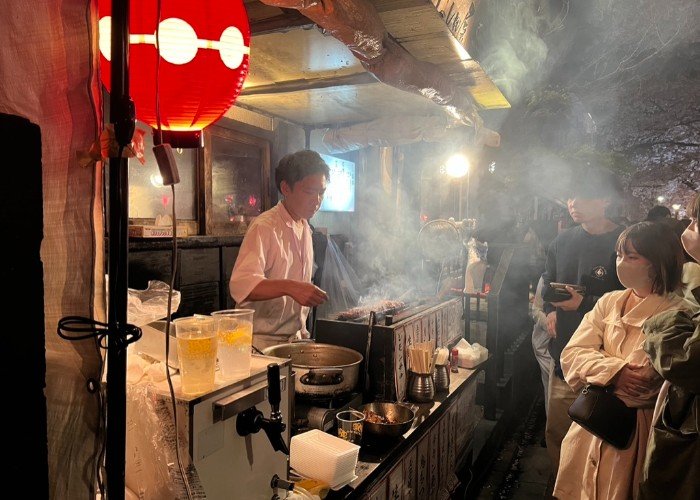 People waiting at a yakitori stand while a man grills chicken skewers