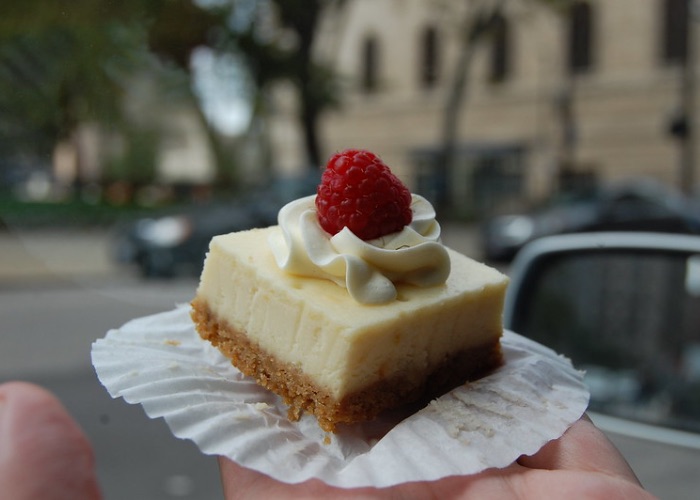 Point of view photo of someone eating a square shaped cheesecake topped with whipped cream and a single raspberry