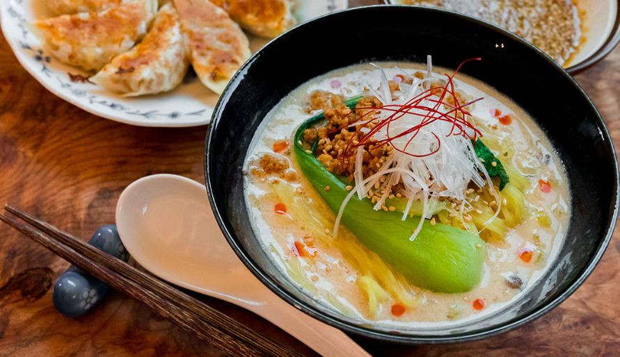 A bowl of vegan ramen on the table with a plate of vegan gyoza behind it