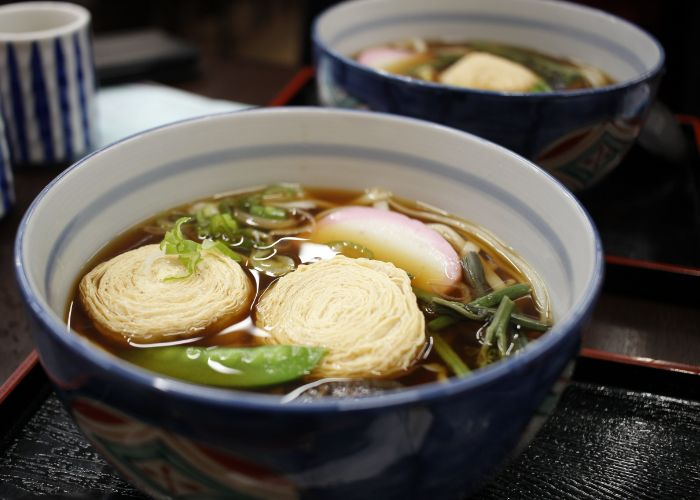 A bowl of yuba soba, featuring a bowl of soba with two rolls of yuba (dried tofu skin) on top and a selection of seasonal vegetables.