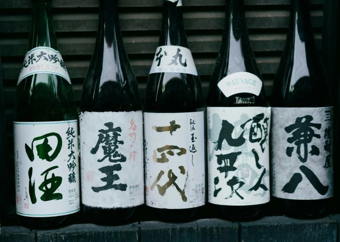 Five bottles of sake lined up on a stone wall. On each, a label with the brand names, written in calligraphy-style kanji.