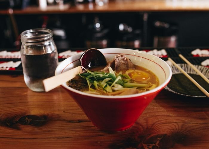 A red bowl of ramen. Floating on top is meat, spring onions, vegetables, and a soft-boiled egg.