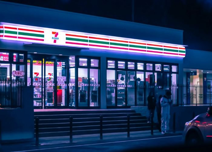 The exterior of a 7-11 in Japan at night, neon lights glowing.