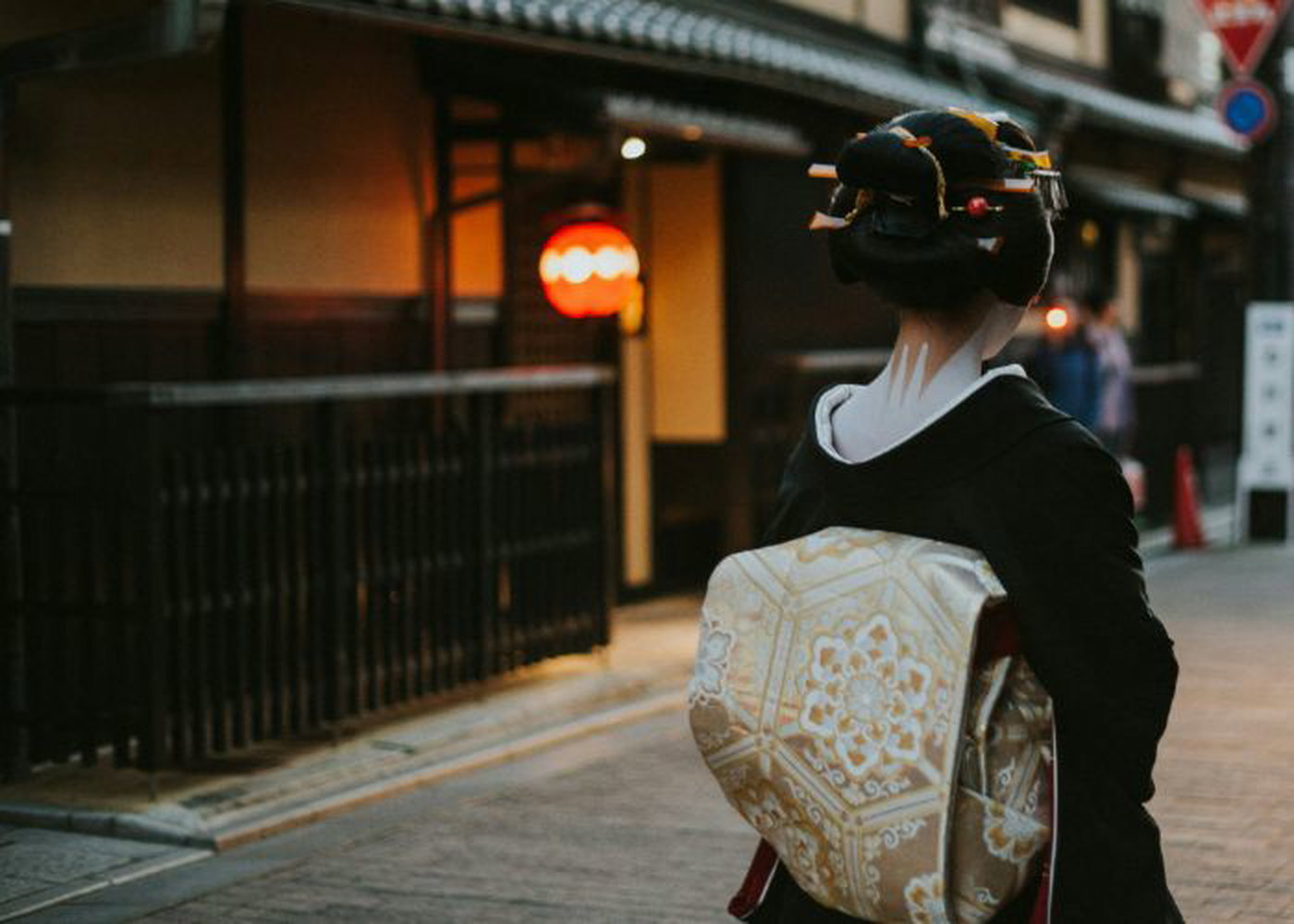 A geisha walks through the streets of Gion. Her hair is up, neck painted, and her golden obi is tied behind her.