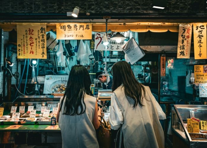 Two girls stand at a shop in Nishiki Market, deciding what to buy.