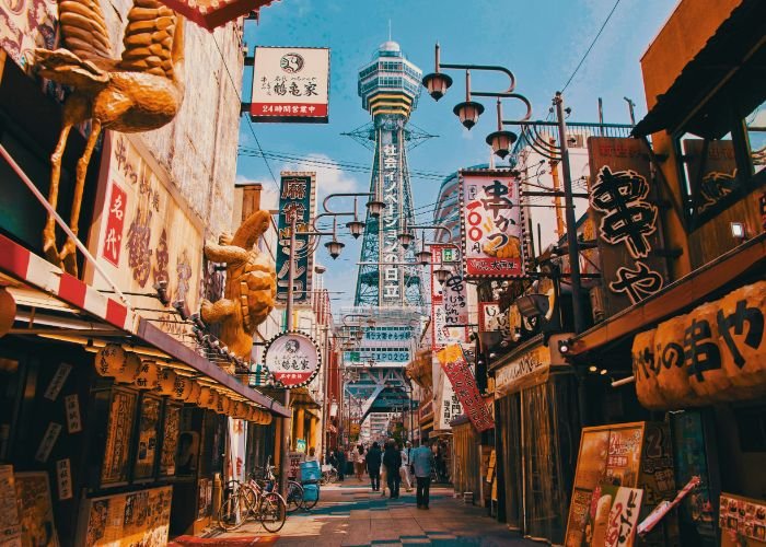 The streets of downtown Osaka, looking up at Tsutenkaku Tower against a blue sky.