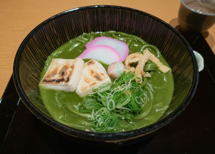 A bowl of noodles, containing dark green matcha-infused broth, topped with tofu, spring onions, and fish cakes.