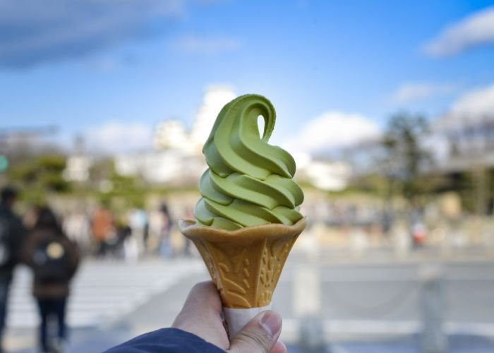 A person holding a matcha ice cream in a wafer cone up in front of them, with the background out of focus.