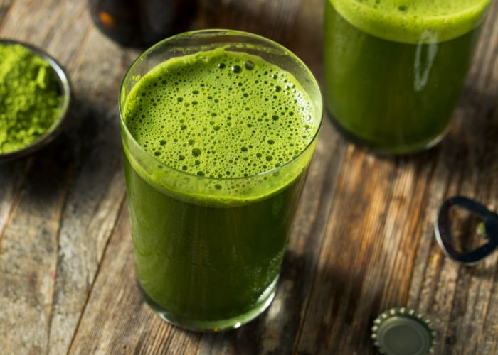 A frothy matcha beer on a dark wood table. Around it, a bottle opening, bottle cap, and matcha powder.