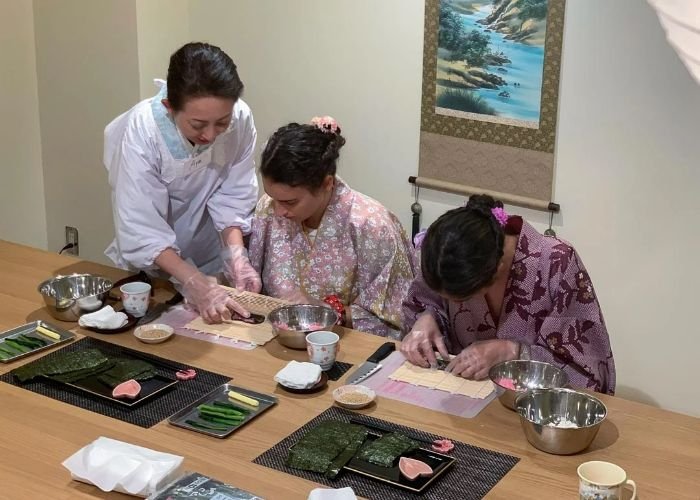 Two ladies in kimono are rolling sushi while a Japanese experts stands, teaching them.