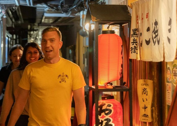 Guests on a bar-hopping food tour in Kyoto smile at the camera, standing outside of a lantern-lit izakaya.