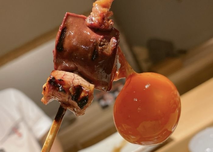 A bright orange yolk is hanging from a skewer, paired with some kind of grilled meat.