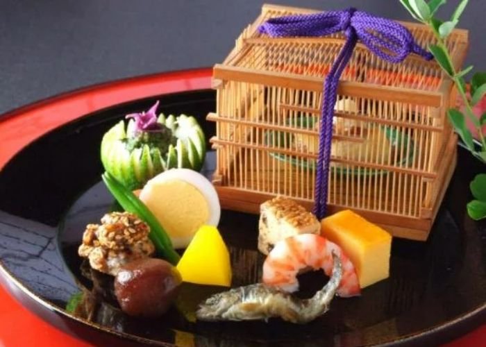 A selection of seasonal vegetables, seafood, and sides are served on a black tray. Behind them, another dish is served in a small bamboo cage with a purple string.