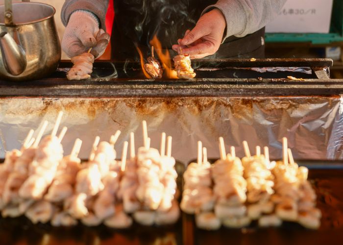 A yakitori vendor grilling chicken skewers and piling them up in the foreground, ready for hungry customers.