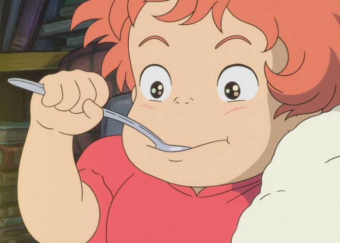 Ponyo sticking a spoon in her mouth, with her cheeks flushed in excitement.
