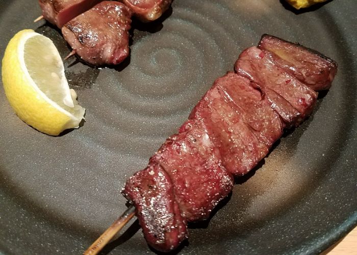 Grilled and skewered meat on a dark grey plate, next to a lemon wedge for flavoring.
