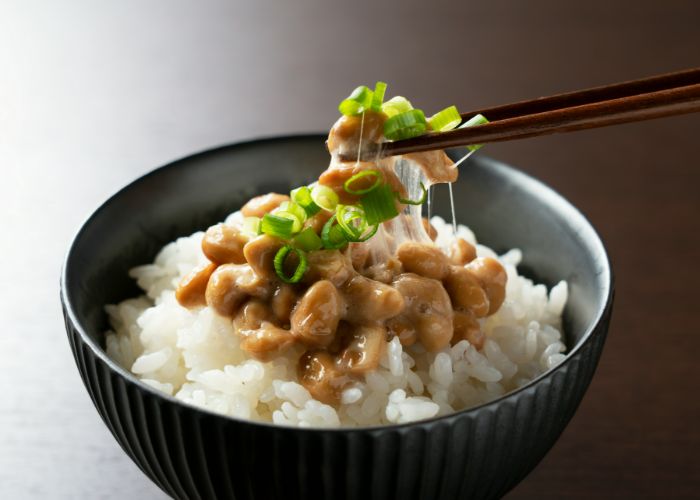 Chopsticks picking up a mouthful from a serving of natto, rice, and spring onions.