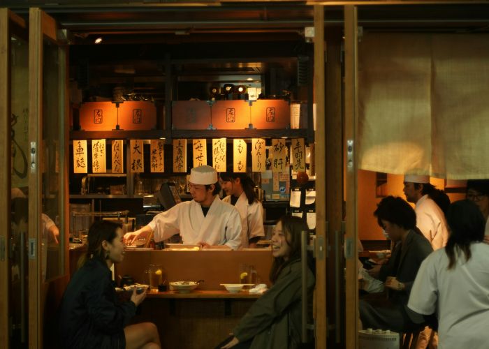 Looking in at a warmly lit izakaya where a chef is cooking while two friends catch up at the counter.