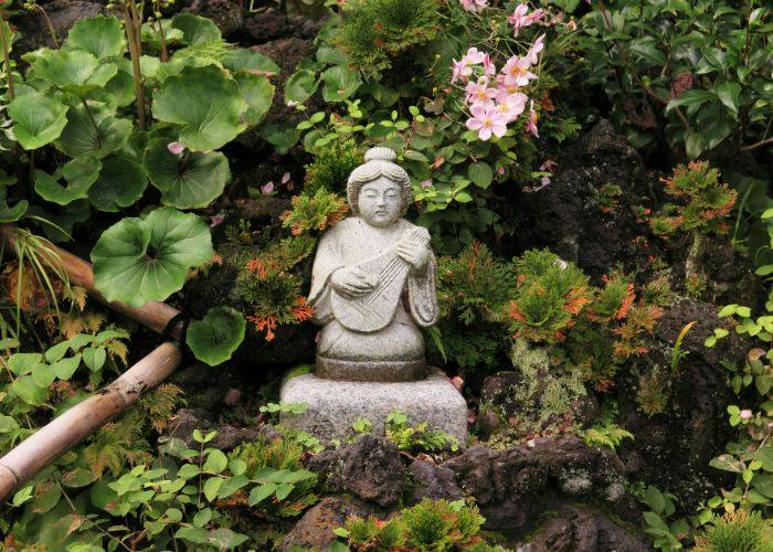 A stone statue of Benzaiten holding a lute, surrounded by Japanese plants with pink flowers.