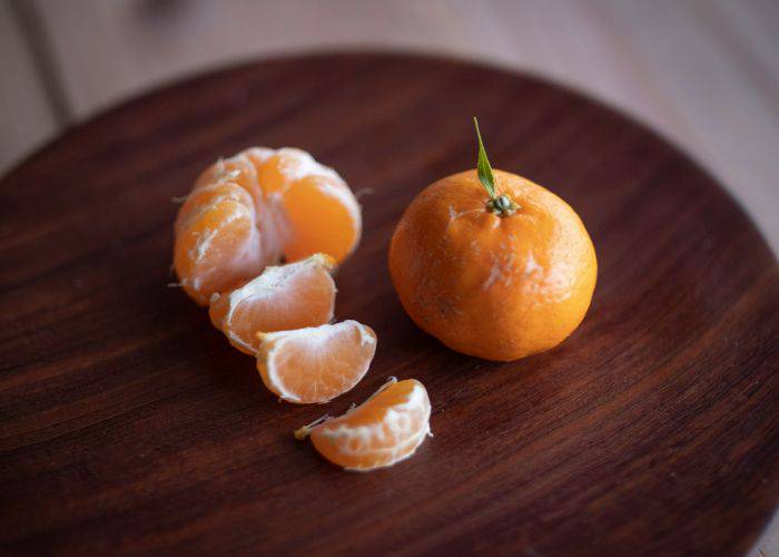 A peeled mikan orange with slices laid out next to another mikan, not yet peeled with a leaf coming off its stem on top of a wooden plate.