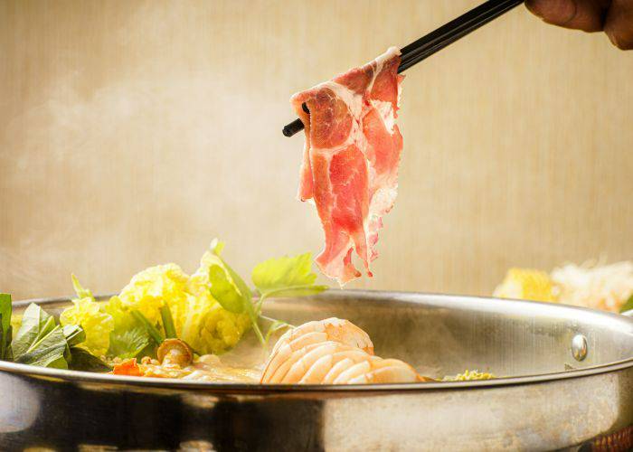 Meats and fresh vegetables cooking in a flavorful hot pot broth. Chopsticks are dunking a slice of meat into it.