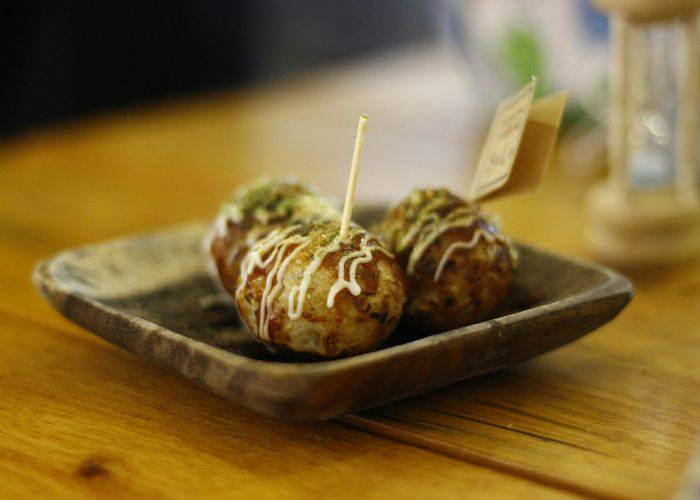 A serving of takoyaki on a ceramic serving tray. A toothpick is poked into the closest takoyaki.