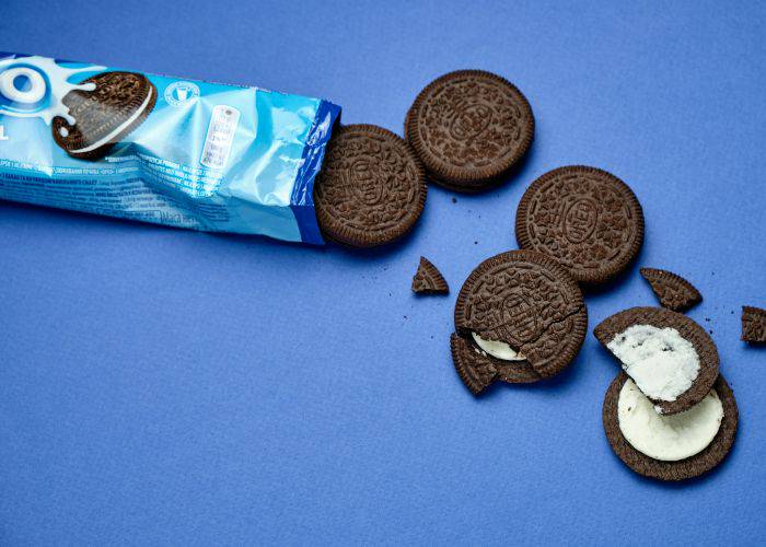 An open package of Oreos spilling out onto a blue background. Some biscuits are broken, revealing the cream inside.