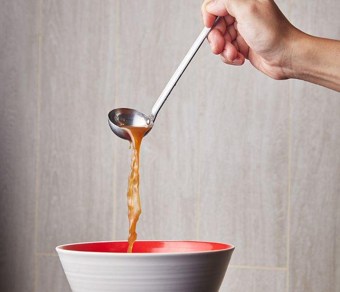 Hand holding a ladle full of hot soup broth, pouring soup into a ramen bowl