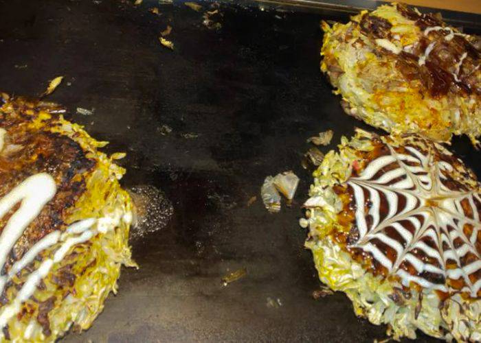 Multiple okonomiyaki on a grill, each smothered in mayo and okonomiyaki sauce.