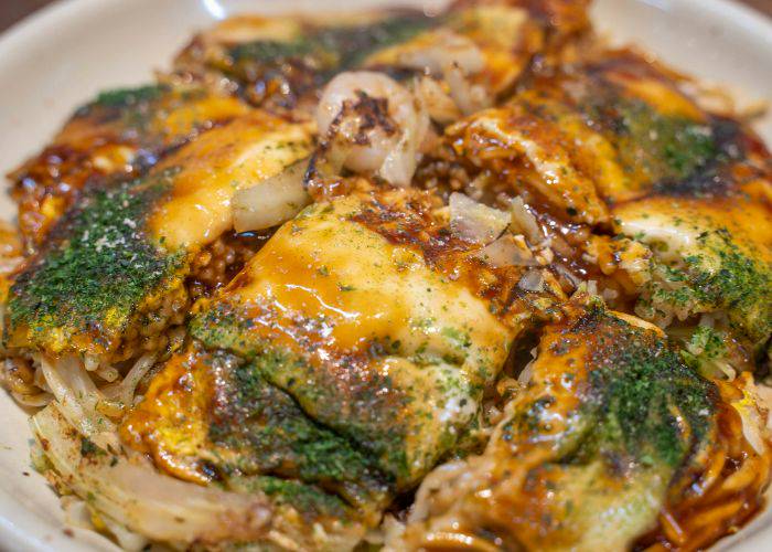 A close-up shot of okonomiyaki, smothered in sauce and garnished with bonito flakes and seaweed.