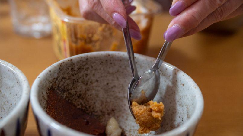 A person with pink nails scraping some miso paste from a spoon and into a bowl.